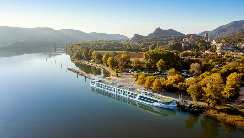Luxury river cruise ship on the Rhône River in France, docked near a wooded area, with a town in the background