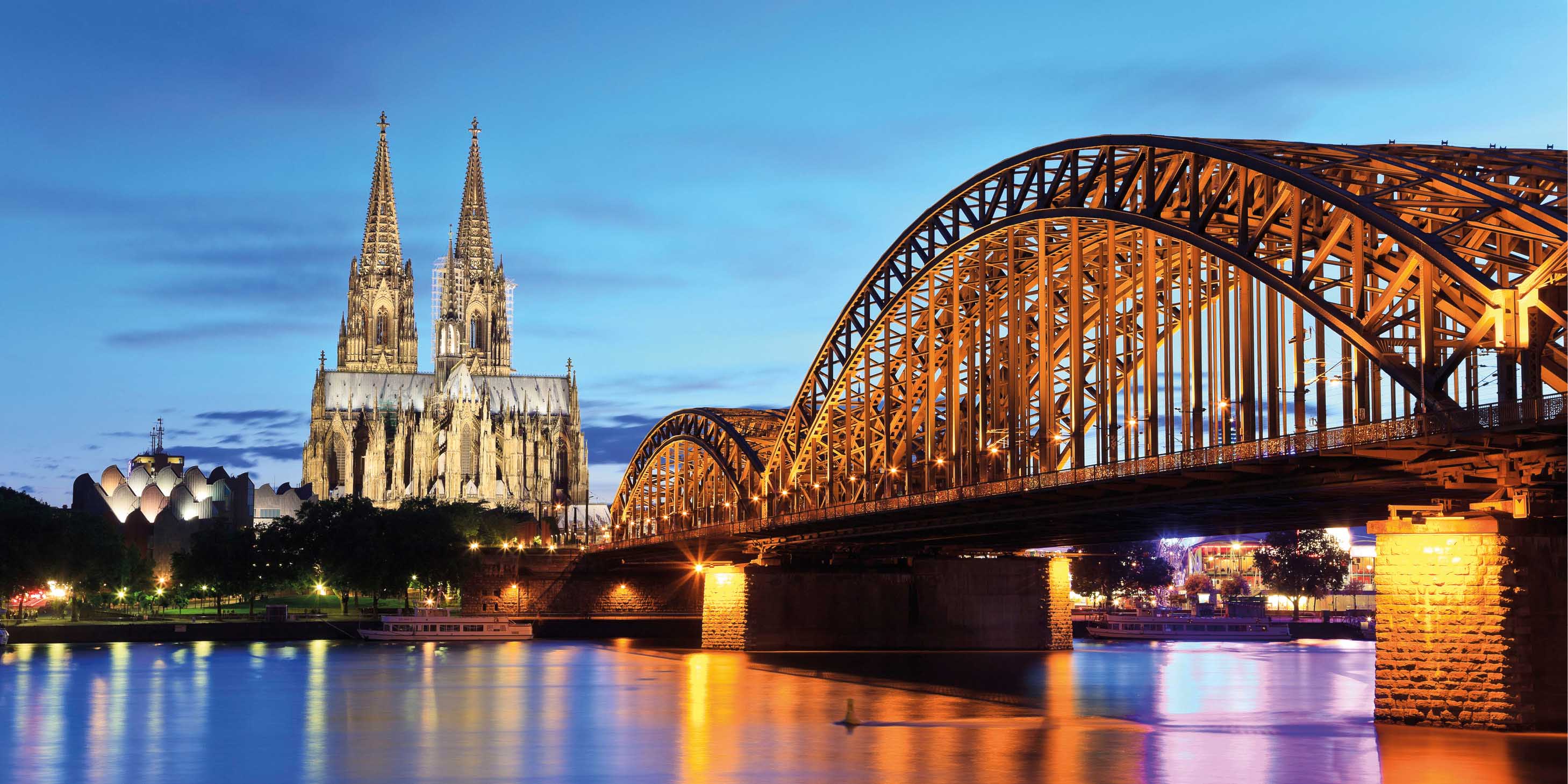 Cologne Cathedral at night with the city glow reflected in the Rhine River and Hohenzollern Bridge