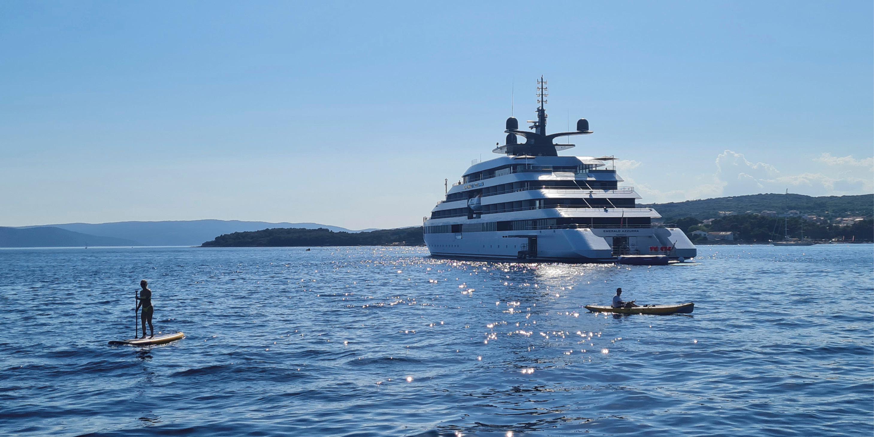 A paddleboarder and kayaker moving across the blue water near a luxury yacht on a sunny day