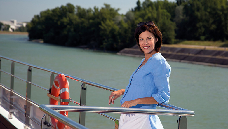 A woman smiling on the top deck of a cruise ship, leaning against the rails, admiring the trees and river