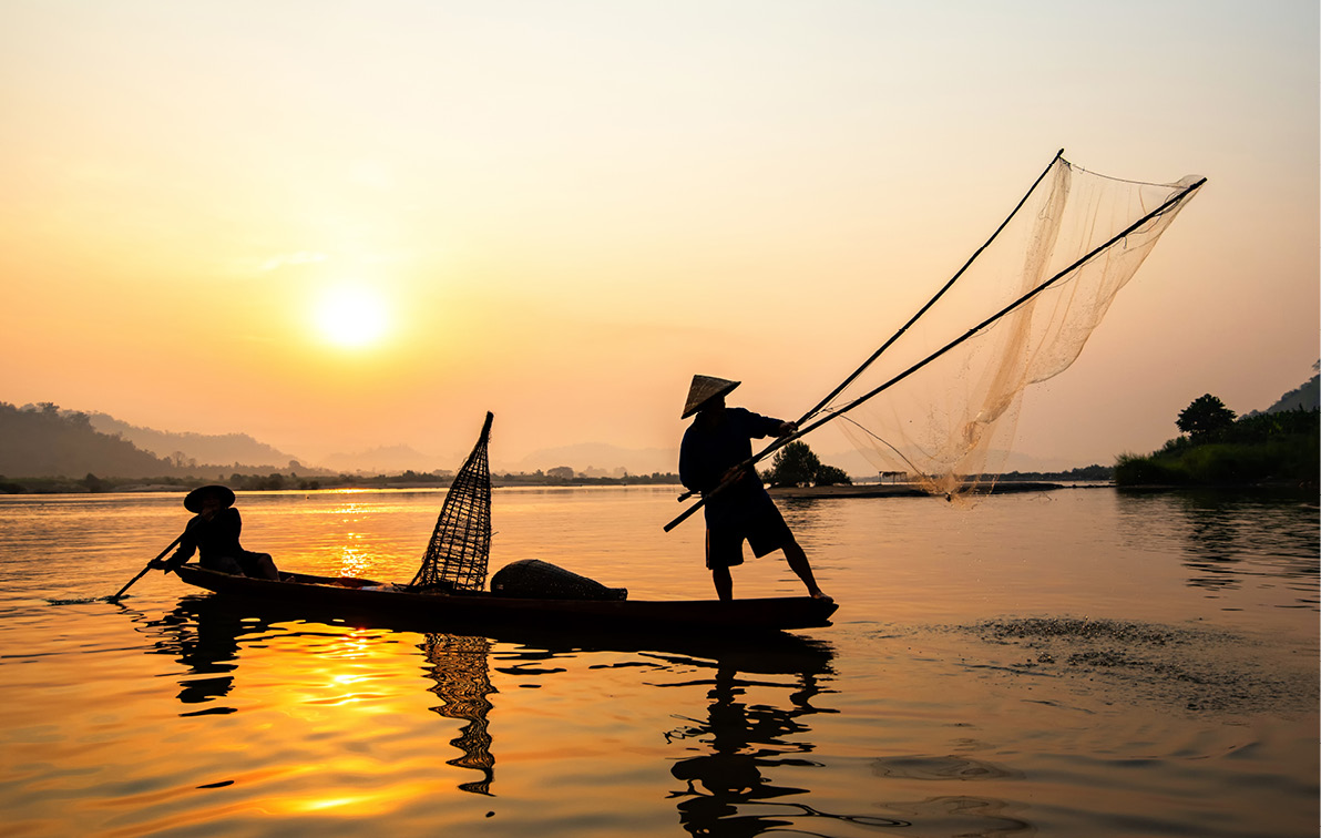 Silhouettes of two men casting nets on authentic Southeast Asia fishing boats set against an orange sunset. 