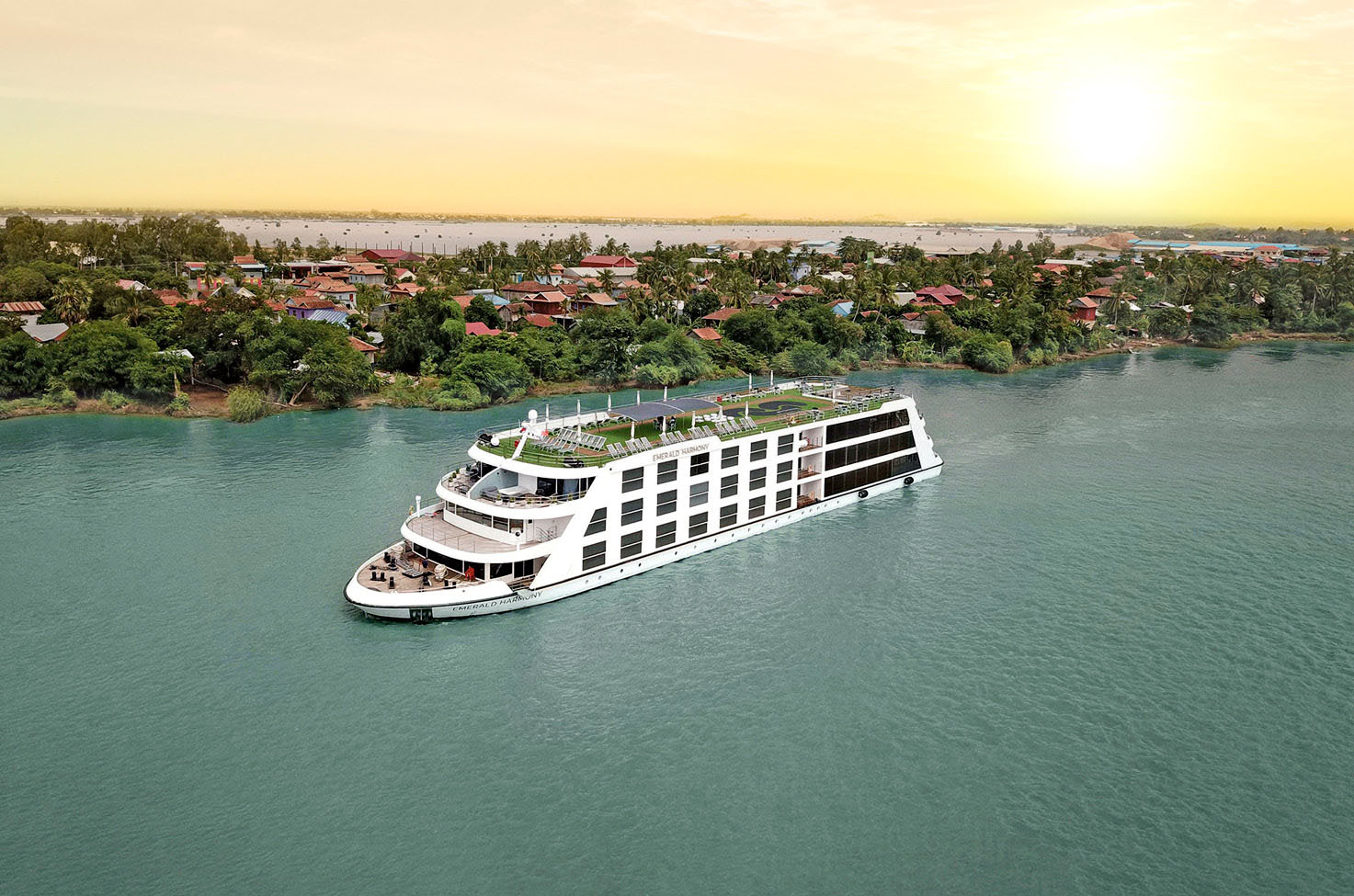 Luxury river cruise ship sailing Mekong River at sunset past land filled with trees and traditional housing