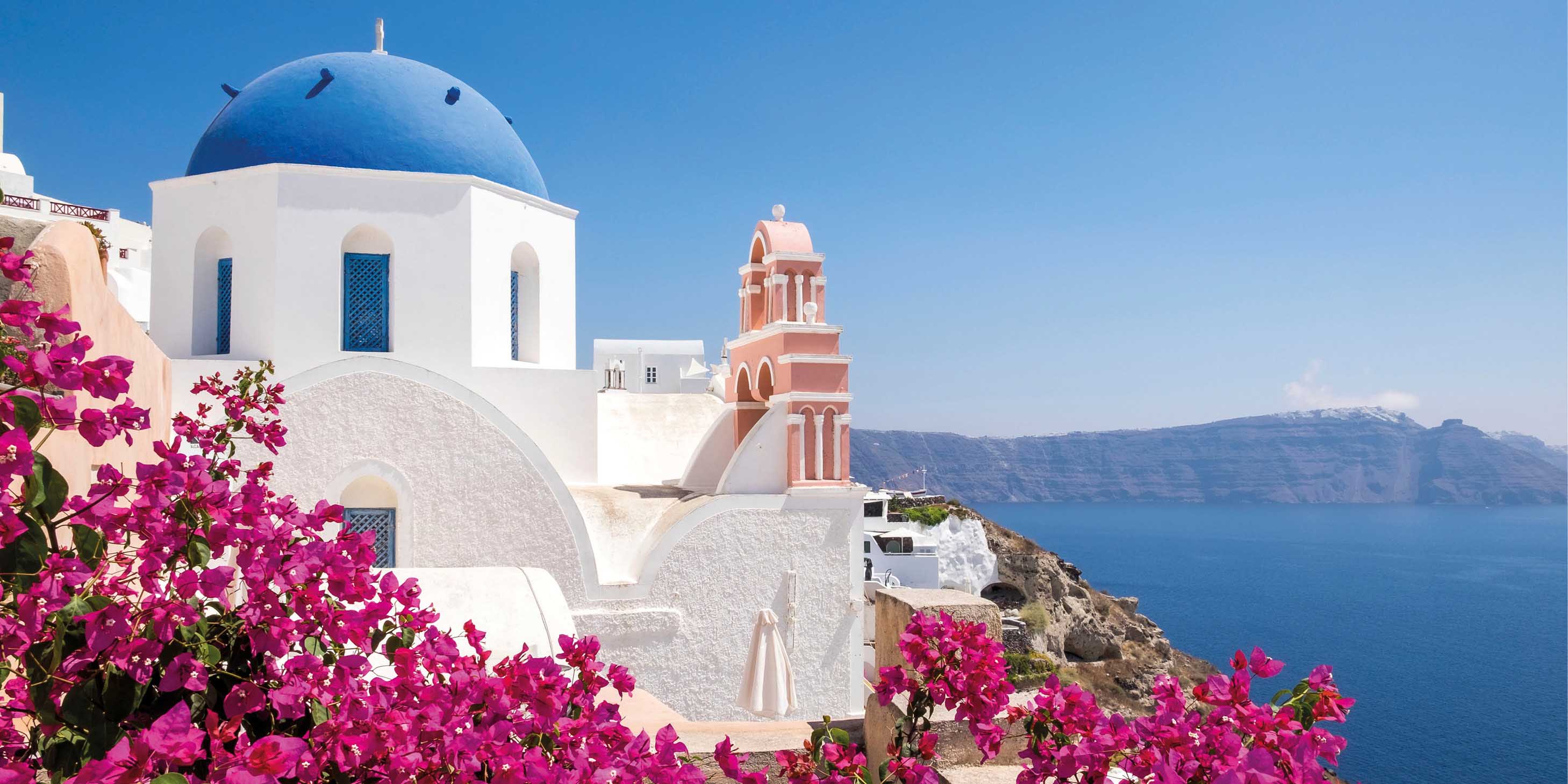 Blue-domed church of Santorini on a bright, sunny day, with the water and sky in background, and flowers in foreground
