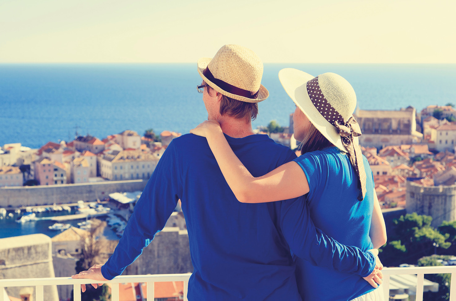 A couple wearing blue shirts and sun hats, as they look over a balcony to view the town and sea below