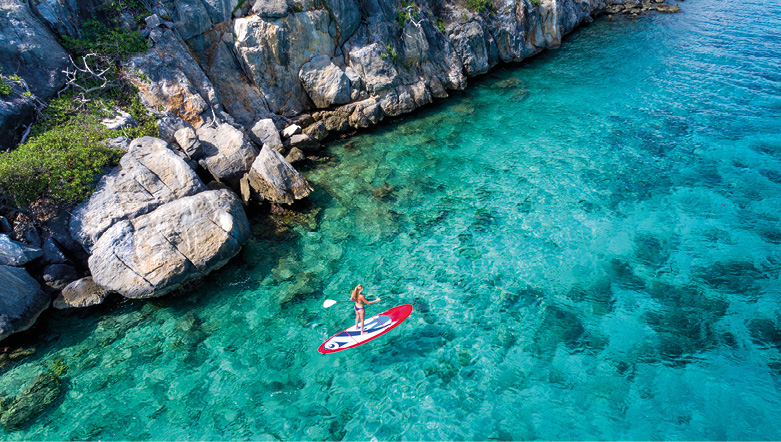 Aerial view of a woman enjoying paddleboarding on the clear blue waters of the Caribbean Sea along the rocky coastline