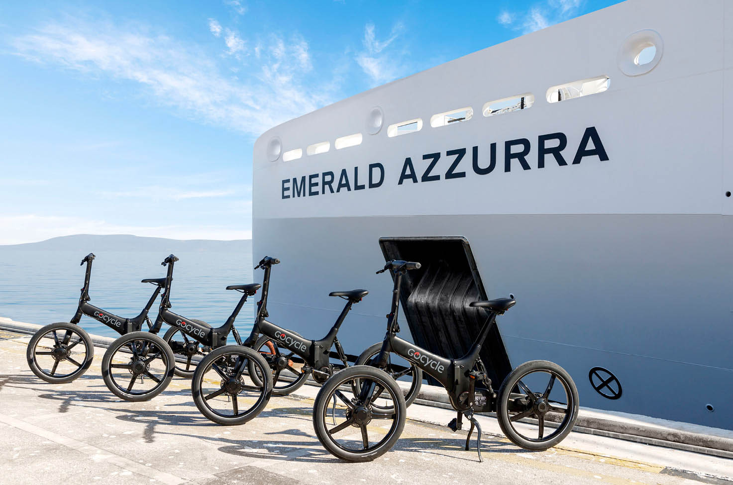 Four electric bikes lined up in front of a luxury yacht, with the blue, light cloudy sky above