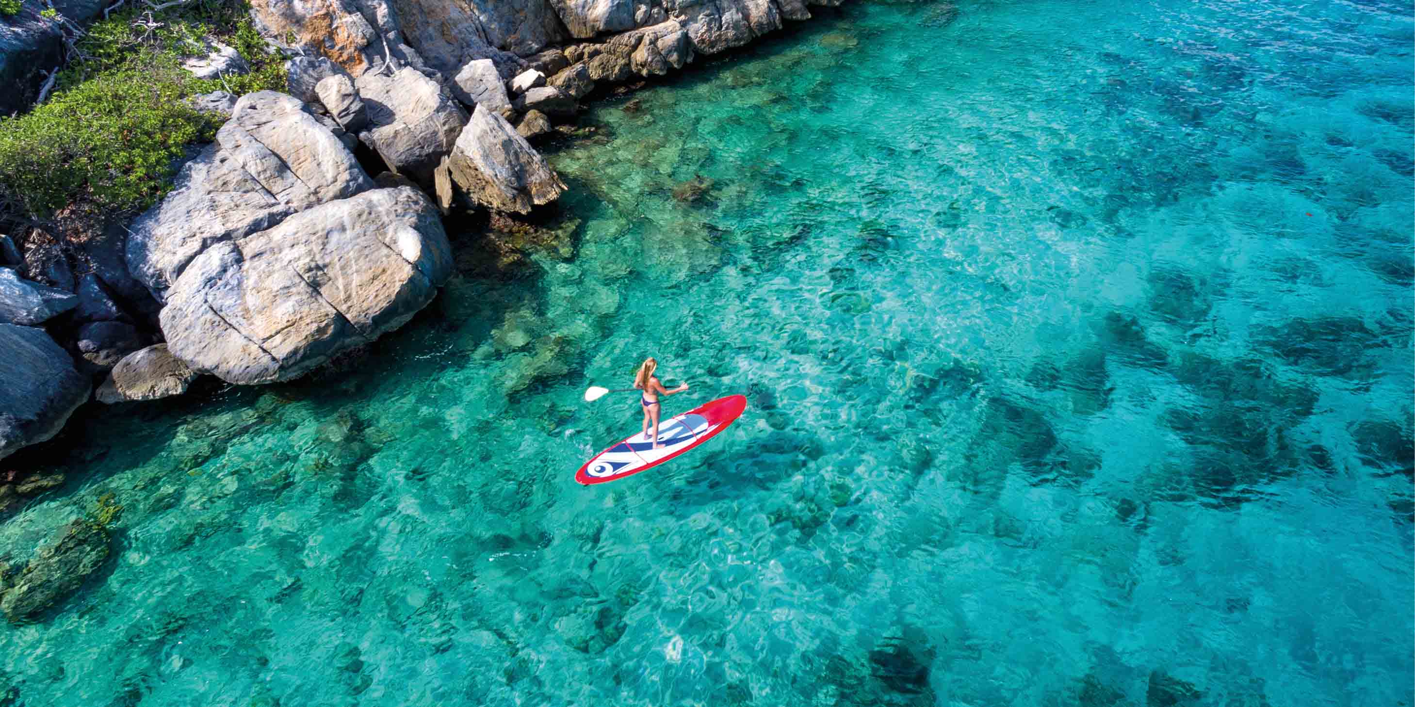 Aerial view of a woman enjoying paddleboarding on the clear blue waters of the Caribbean Sea along the rocky coastline
